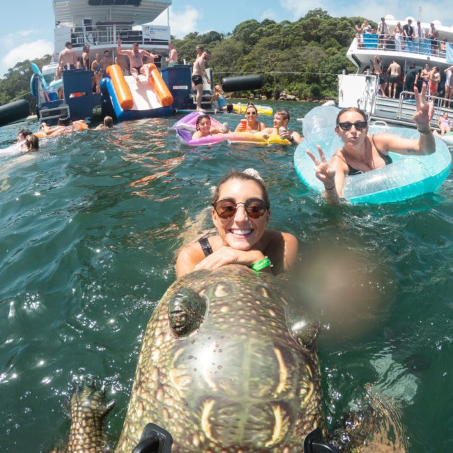 People swimming and floating on inflatables in a lake near a luxury yacht with water slides. One person in the foreground is holding an inflatable crocodile. Other people are seen on the yacht and slides, making it a lively Catamaran party Sydney event.