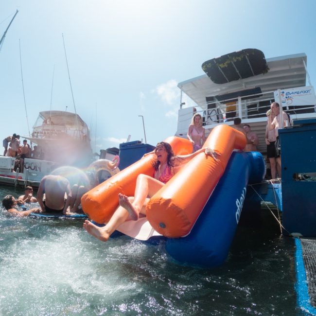 Person sliding down an orange inflatable slide into the water from a boat, surrounded by people on other boats and in the water. Bright, sunny day with clear blue sky—perfect for a catamaran party in Sydney or a corporate boat event at Sydney Harbour.