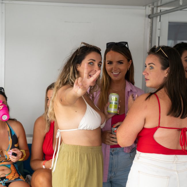 A group of women socializing indoors, one woman in a white bikini top and green skirt gesturing a peace sign towards the camera, while others chat and hold drinks. The atmosphere is reminiscent of a lively catamaran party in Sydney.