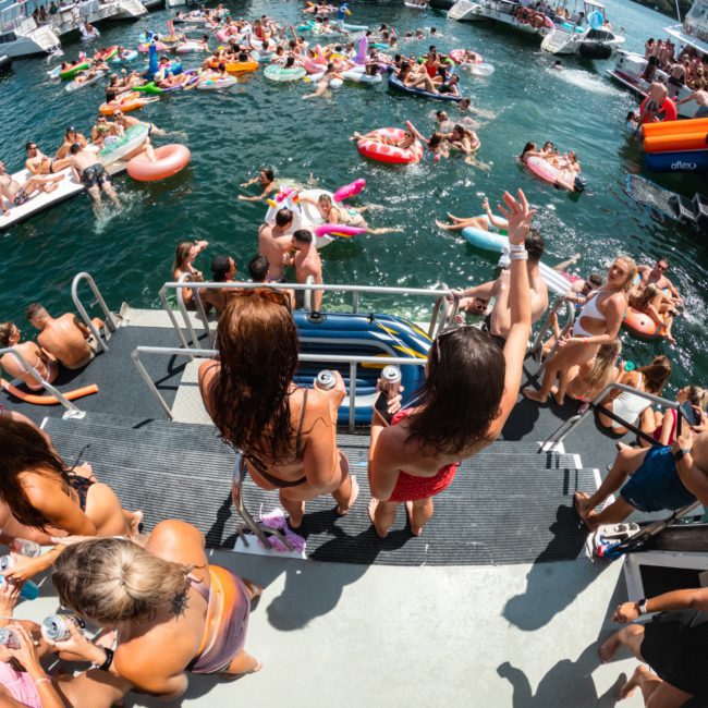 A group of people on boats and inflatable floats are enjoying a lively gathering in the water, with some standing on a platform, holding drinks, while others swim or lounge. The scene features a luxury yacht hire Sydney experience that adds an extra touch of elegance to the celebration.