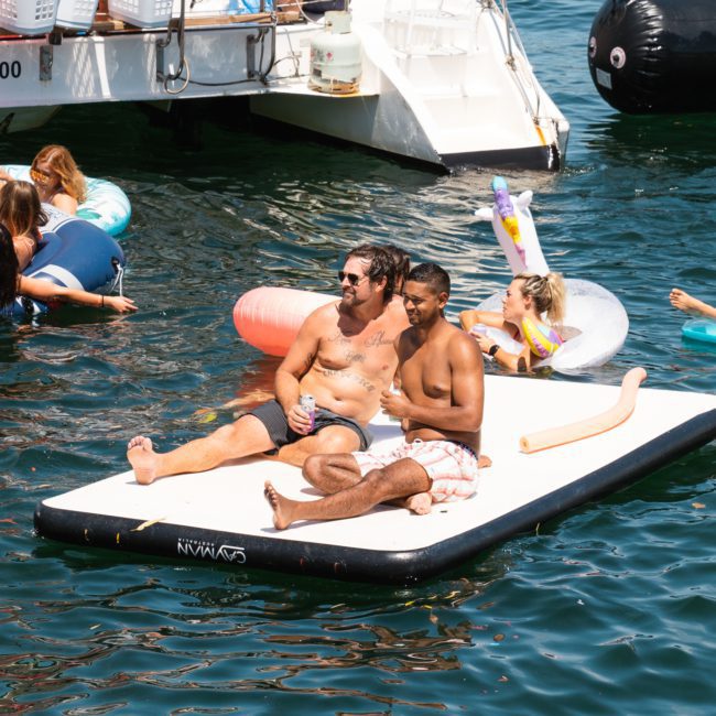 Two people are sitting on an inflated float in the water, surrounded by others swimming and relaxing on various inflatable devices near a catamaran during a Sydney boat party hire event.