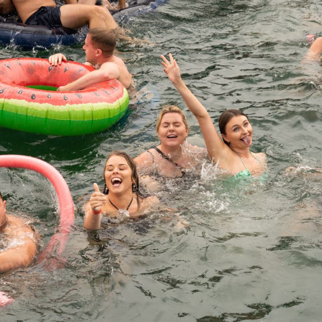 People in a body of water enjoying a social gathering with inflatable tubes and pool noodles, against the backdrop of a private yacht charter in Sydney Harbour. Some are smiling and making gestures, creating a lively and joyful atmosphere.