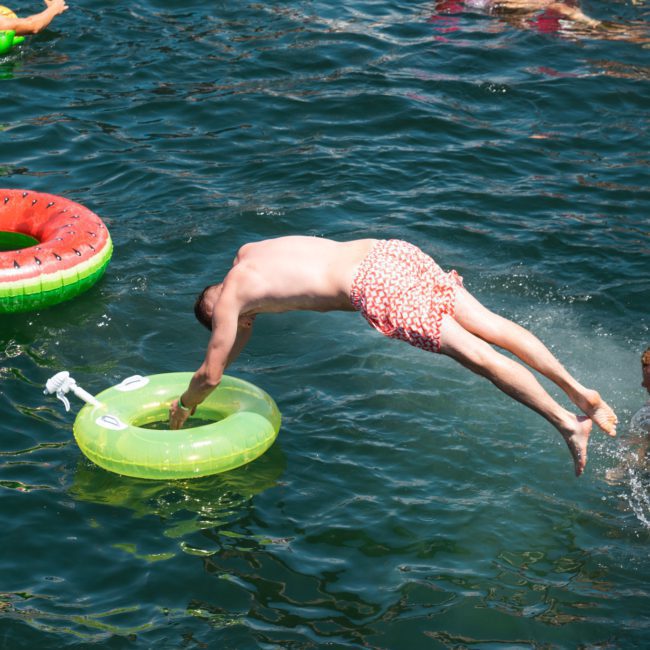 A person in red swim trunks dives into clear water near a green inflatable ring, while another holds their head, partially submerged. Two more inflatables float nearby on the serene waters of a luxury yacht hire Sydney Harbour, offering fun and relaxation amidst breathtaking views.