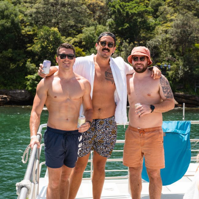 Three men with drinks in hand stand together on a boat in sunny weather. The man in the middle wears a white shirt and printed shorts, while the other two are shirtless and wear shorts. Trees and water are in the background, setting the perfect scene for a catamaran party Sydney style.