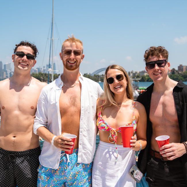 Four people pose for a photo on a boat, each holding a red cup. The scene includes other boats, water, and distant buildings. Enjoying a Sydney boat party hire, they capture the essence of fun on the water.