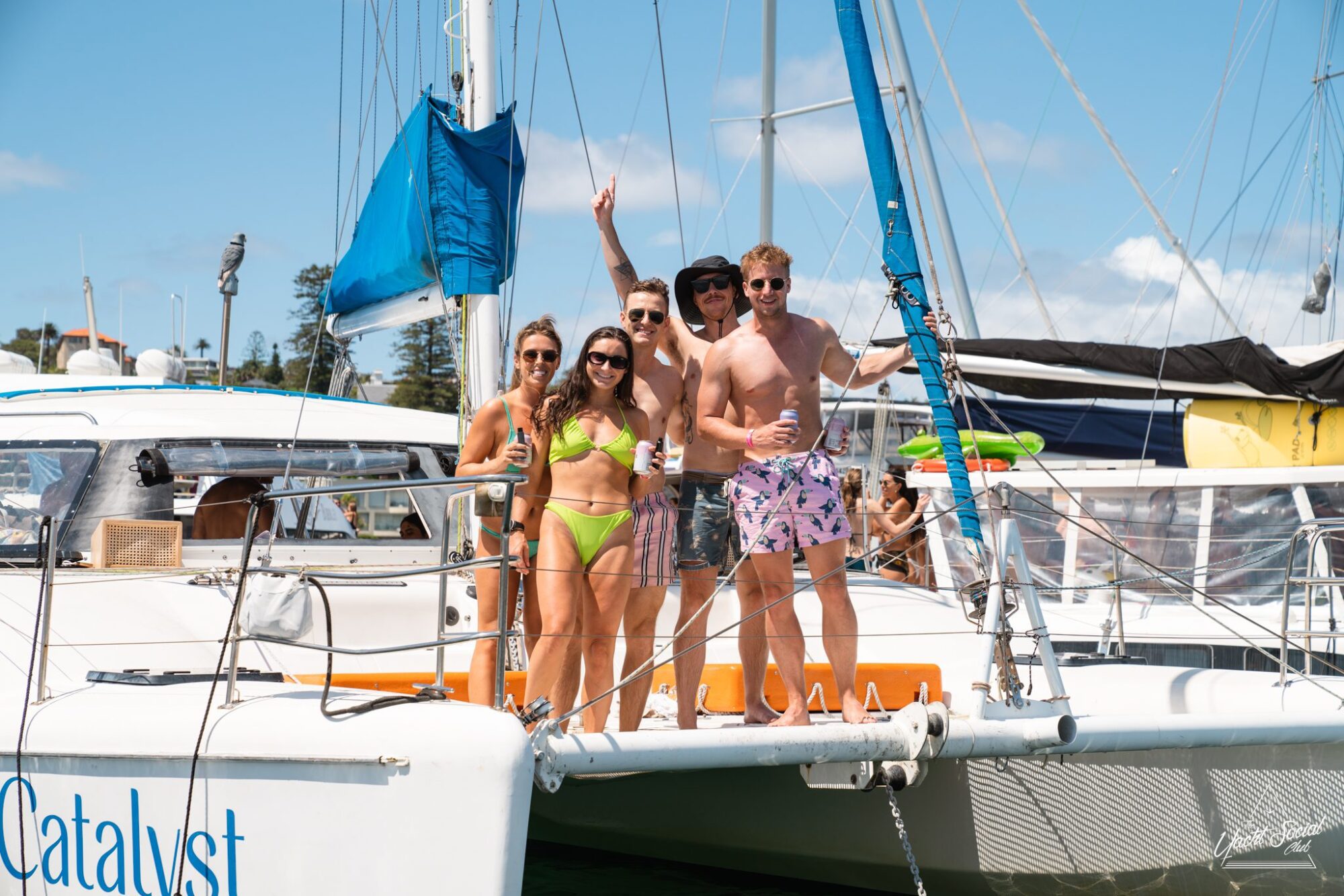 A group of five people stand on the deck of a sailboat named "Catalyst," smiling and posing for the camera. They are in casual, summery attire, enjoying a sunny day. Perfect for a catamaran party Sydney or private yacht charter Sydney Harbour adventure.