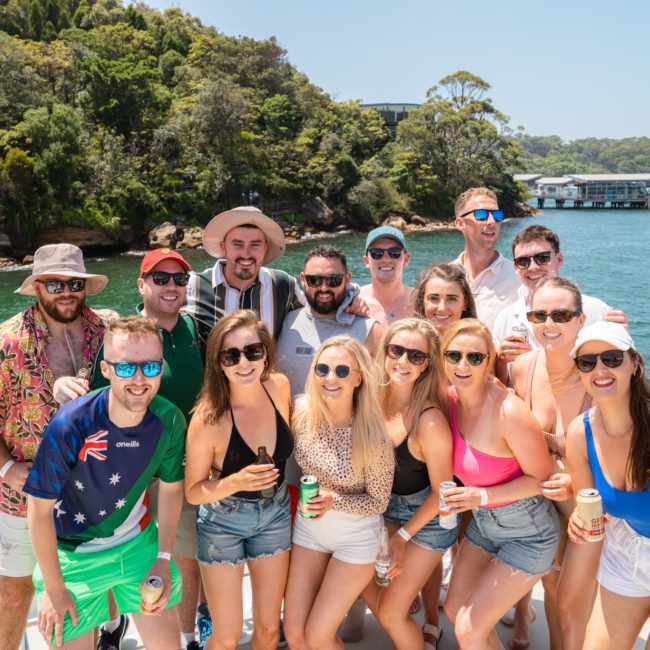 A group of people, casually dressed in summer attire, pose together on a luxury yacht hire in Sydney with a scenic backdrop of greenery and water. Most of them are holding beverages and wearing sunglasses.