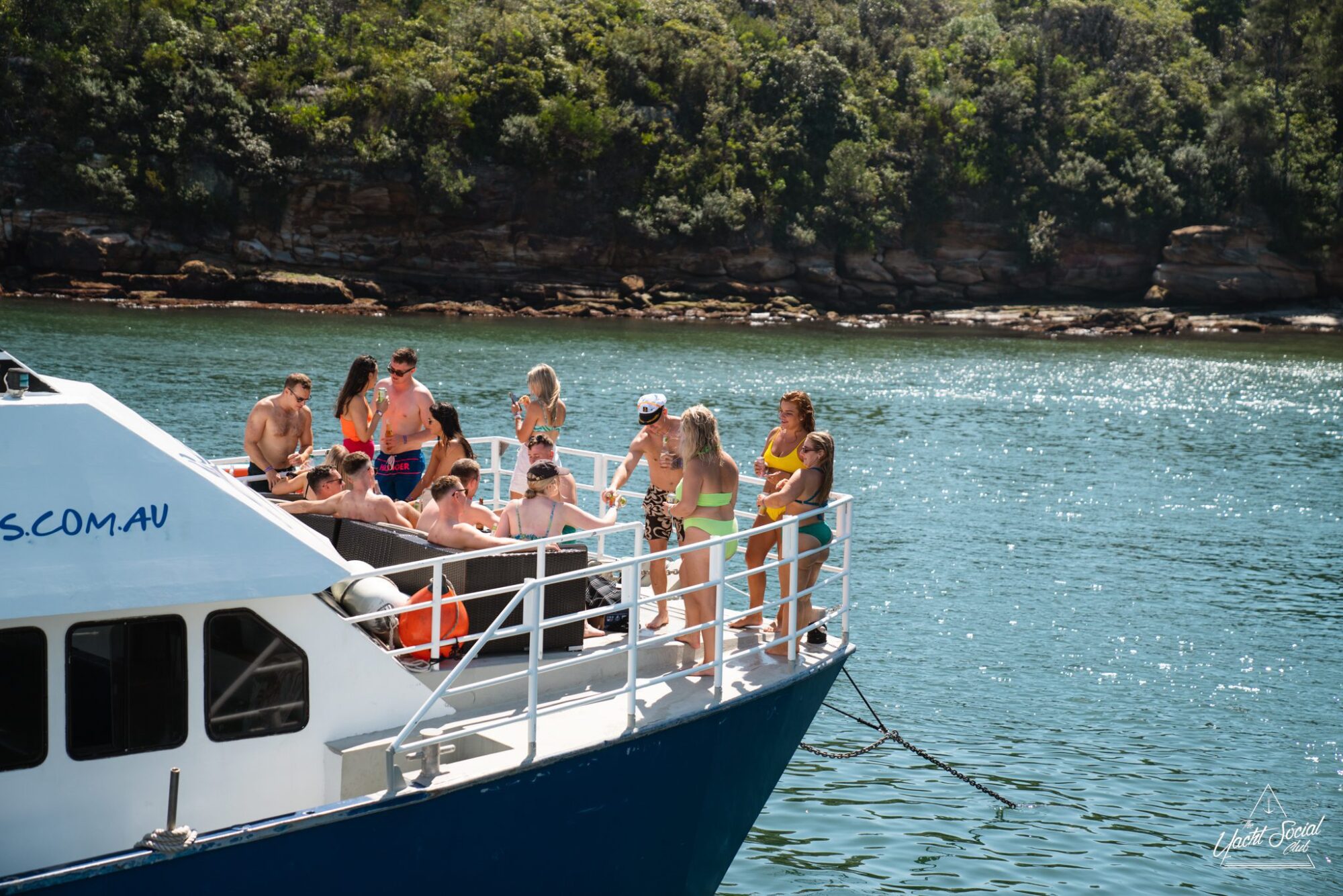 A group of people enjoying a sunny day on a catamaran party in Sydney, docked near a forested shoreline. The boat's white exterior contrasts beautifully with the blue water.