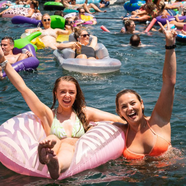 People in swimsuits enjoy floating on inflatable tubes in a large body of water, smiling, and raising their arms in celebration on a sunny day during a Sydney boat party hire.