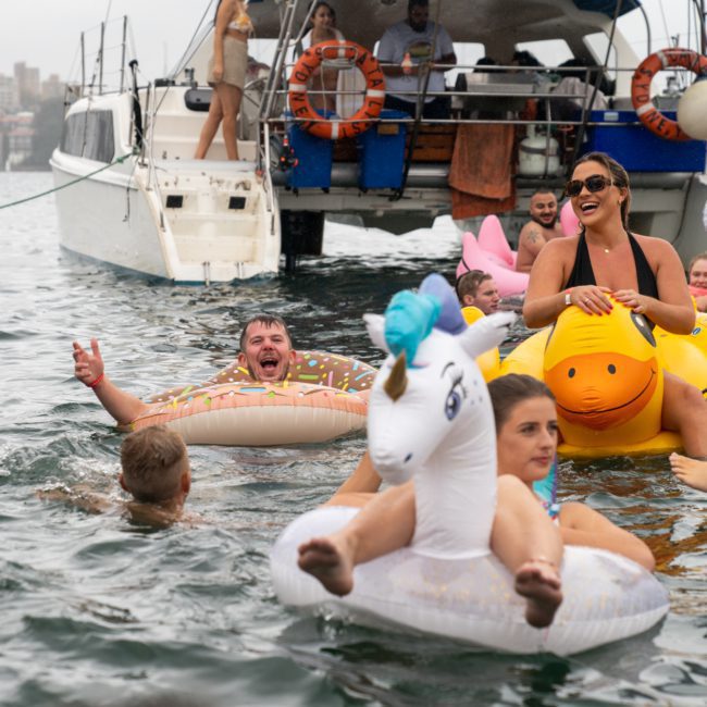 A group of people enjoying a pool party on a luxury yacht with various inflatable floats, including a unicorn and a donut. The background shows city buildings and cloudy skies, highlighting the excitement of DJ boat hire Sydney.