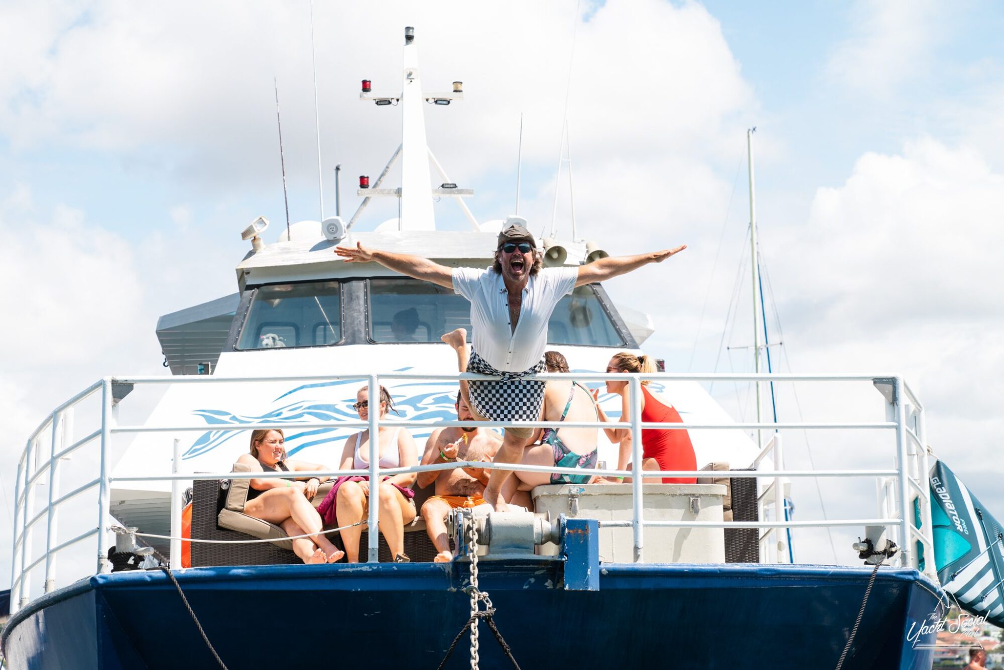 A group of people enjoy lounging at the front of a luxury yacht under sunny skies, while one person stands with arms outstretched in a lively pose.