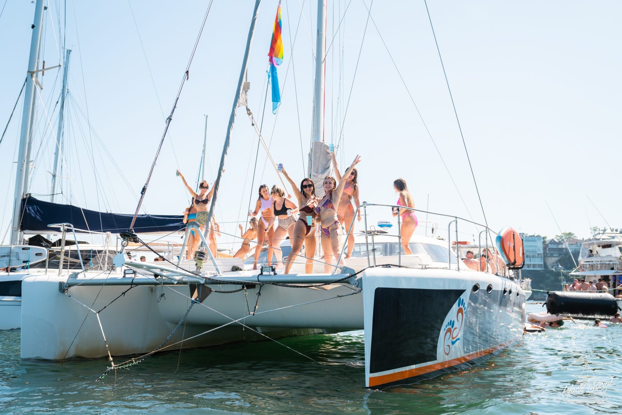 A group of people in swimsuits are standing and posing on the deck of a catamaran sailboat in a marina with other boats visible in the background, enjoying a lively catamaran party in Sydney.