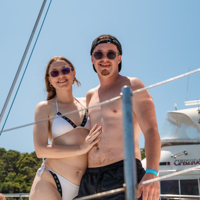 Two people in swimwear stand together on a boat on a sunny day. The woman wears a white bikini, and the man wears black swim trunks and a cap. Both are smiling at their Sydney boat party hire on the sparkling waters.