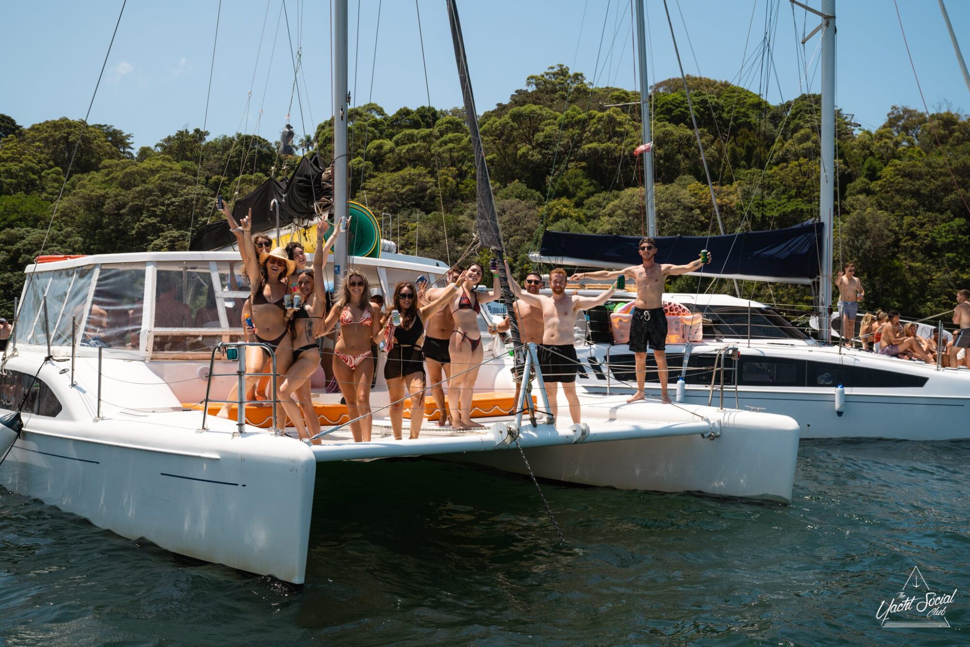 Group of people standing on a white catamaran boat, smiling and raising their arms, with trees and other boats in the background, enjoying a corporate boat event on Sydney Harbour.