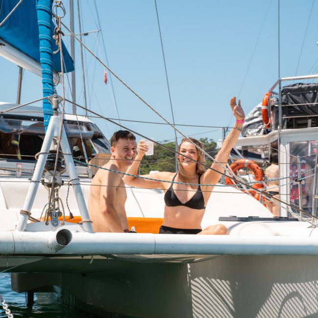 Two people in swimsuits sit and wave on the deck of a private yacht charter in sunny weather, with other boats and a blue sky in the background.