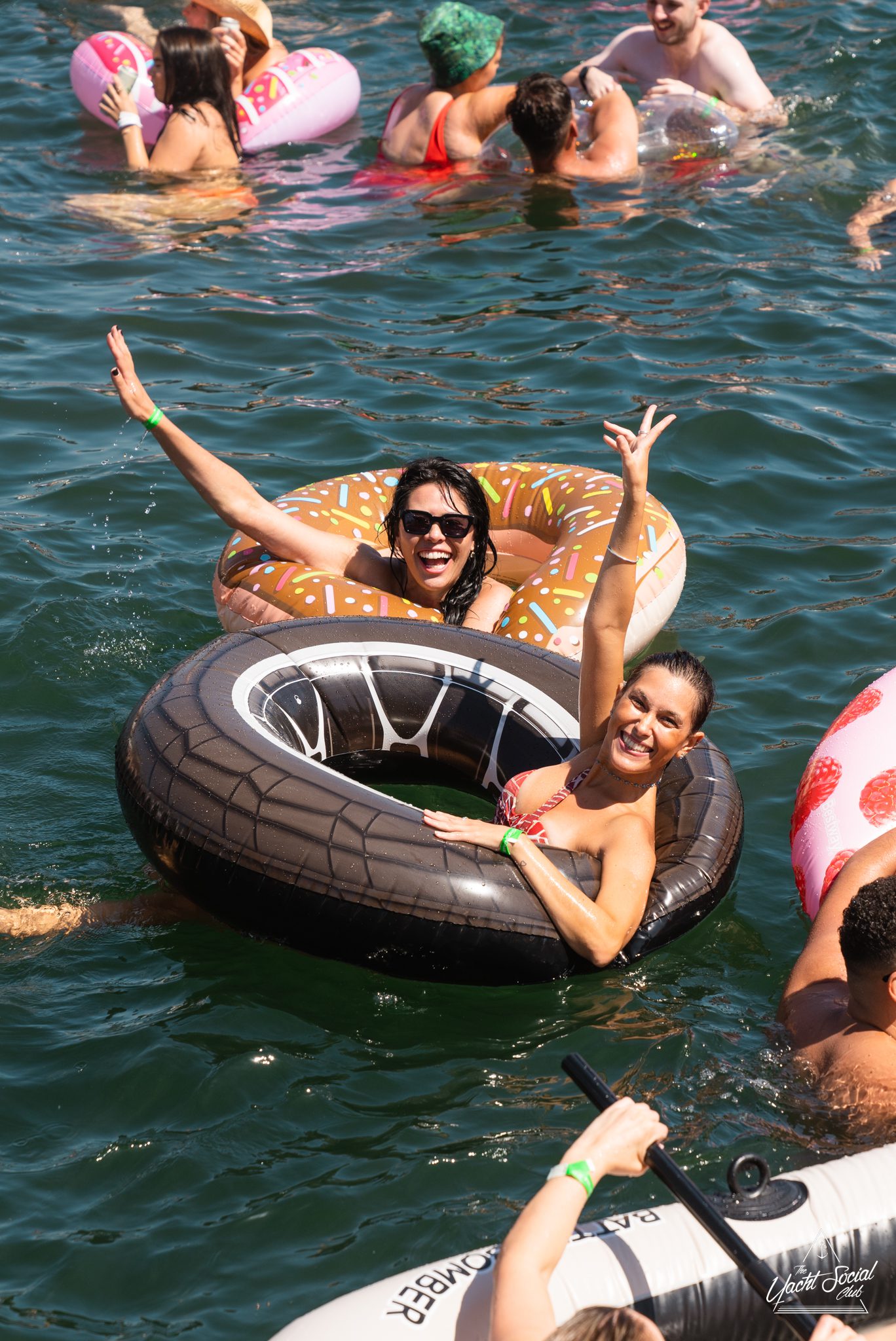 Two women in inflatable pool tubes, one with a donut design and the other resembling a tire, smile and wave while floating in a body of water surrounded by other swimmers. Imagine the fun amplified with DJ boat hire Sydney for an unforgettable experience on the water!