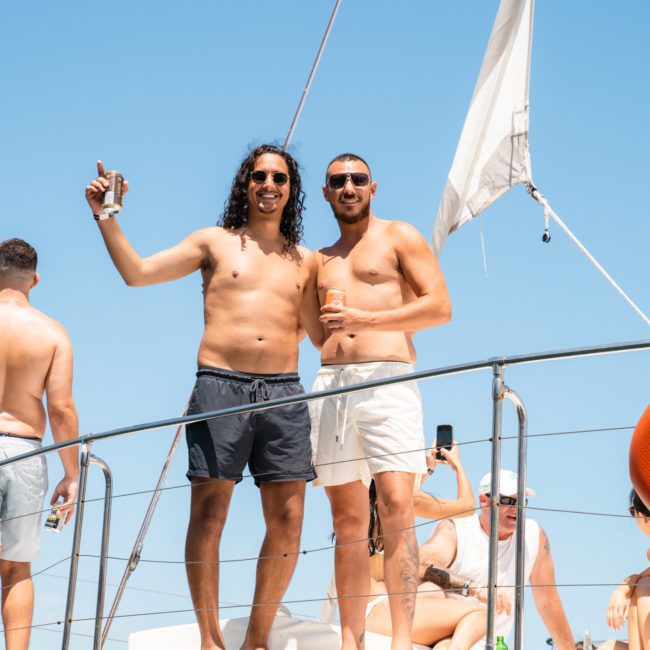 Two men stand side by side on the deck of a boat, smiling. Others are also on the boat, some taking pictures during a Sydney boat party hire. The sky is clear and blue.