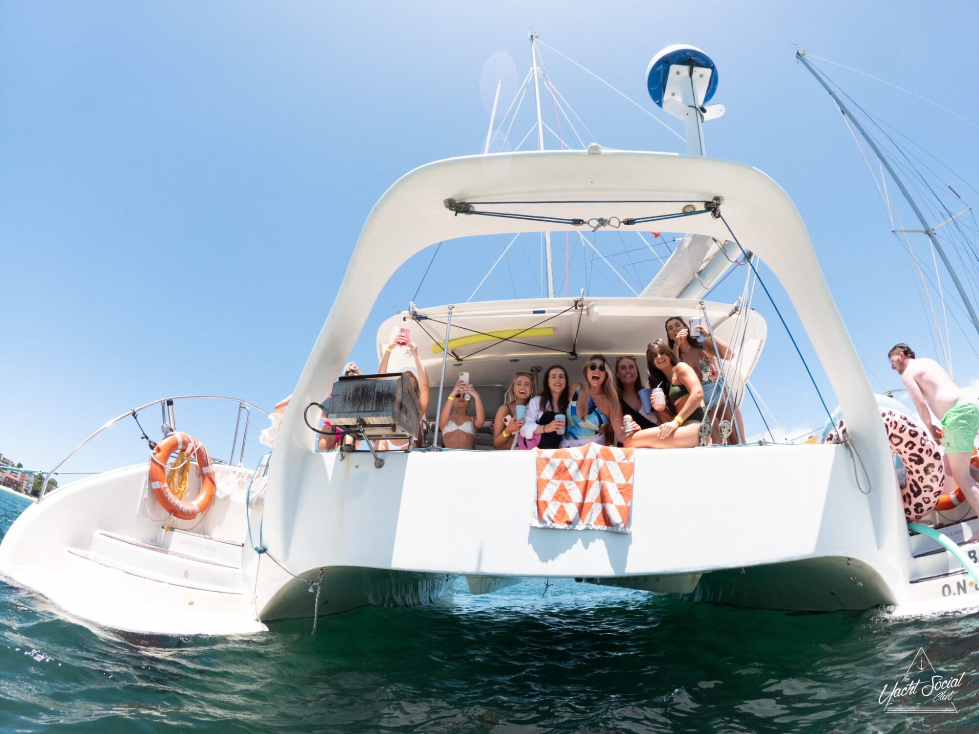 A group of people on a catamaran smiling and posing, with a bright blue sky in the background. The luxury yacht hire Sydney has a striped towel hanging at the back and is floating on clear water.