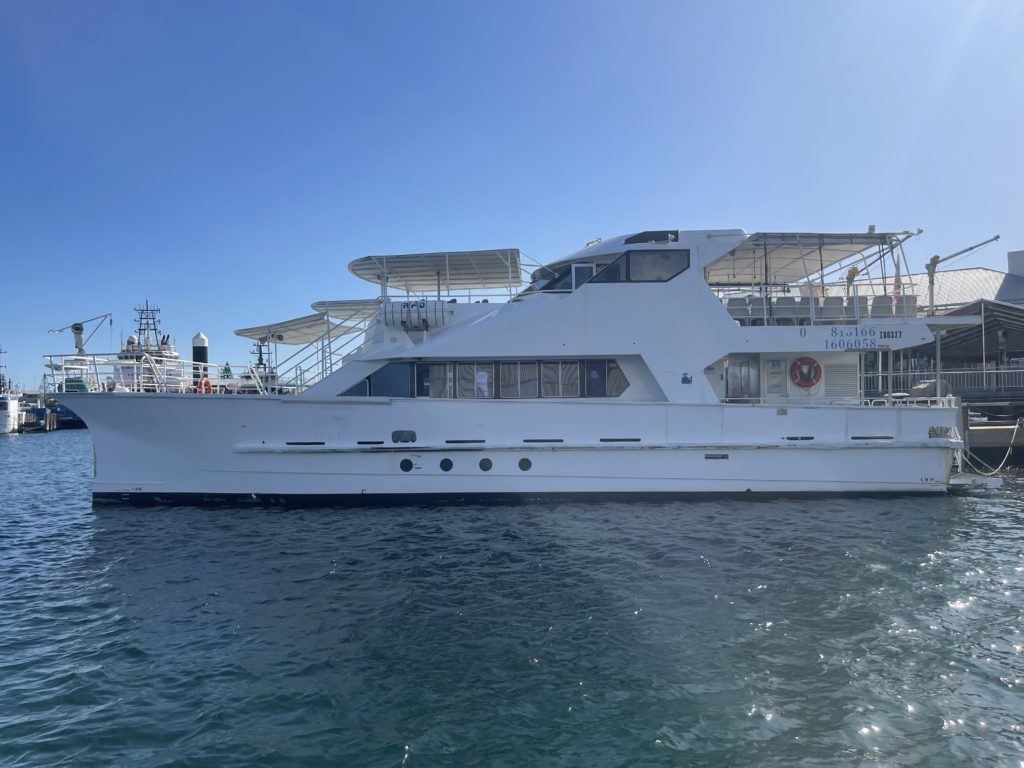 The DJ Boat, a large white yacht with multiple decks and top-mounted solar panels, is docked in calm waters on a sunny day. Ideal for a Catamaran party in Sydney, the sides are adorned with various signs and safety equipment.