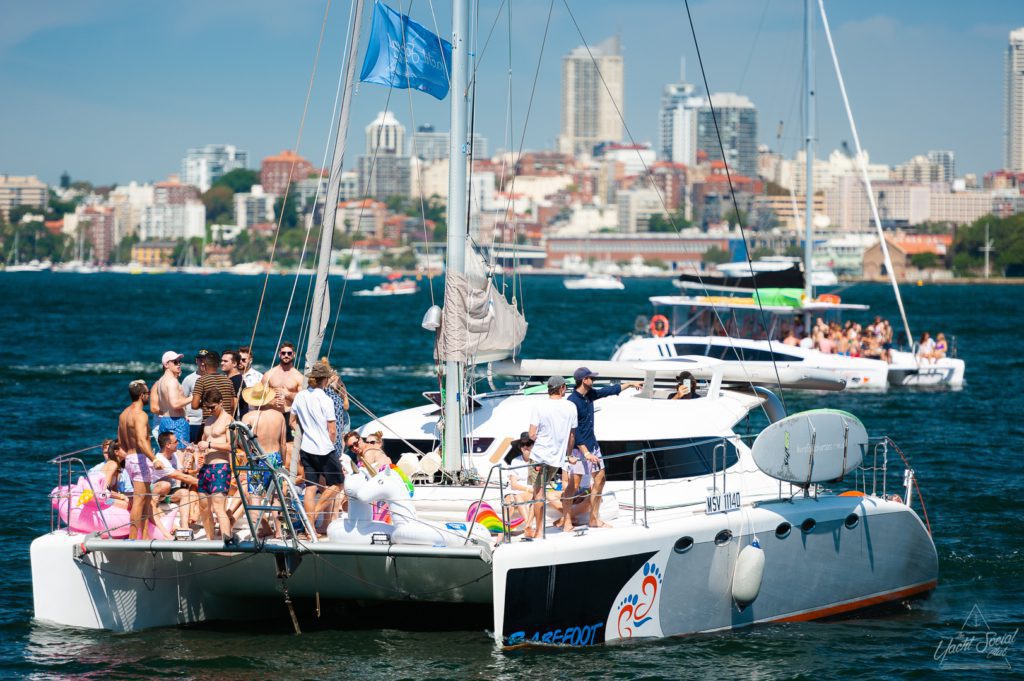 A group of people are gathered on Barefoot, a large private yacht charter in Sydney Harbour, with city buildings in the background. Several other boats are visible on the water, adding to the lively scene.