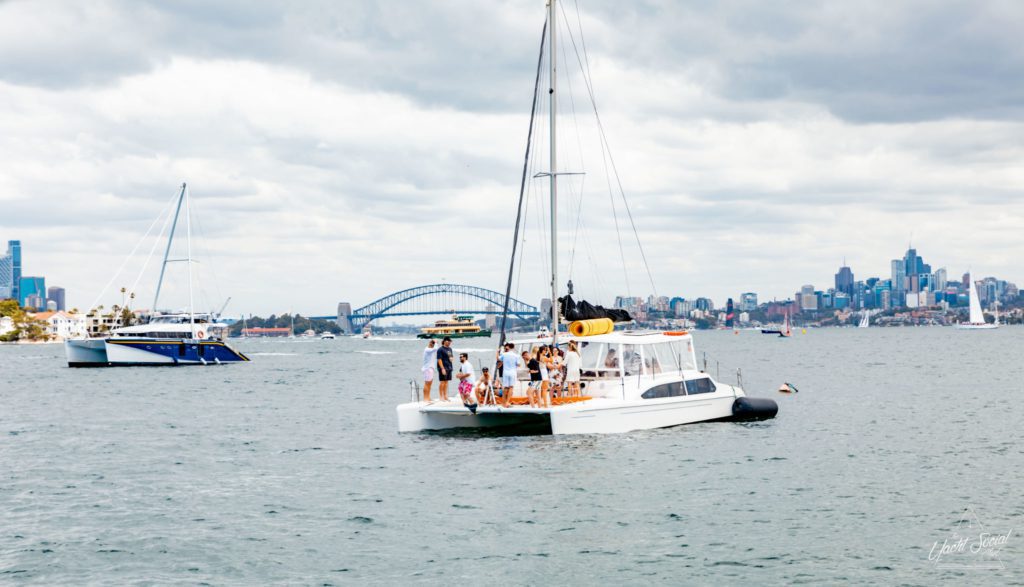 A catamaran with people onboard sails in a bay, perfect for a Zulu party Sydney. In the background, another sailboat and a bridge are visible, with a city skyline under a cloudy sky.