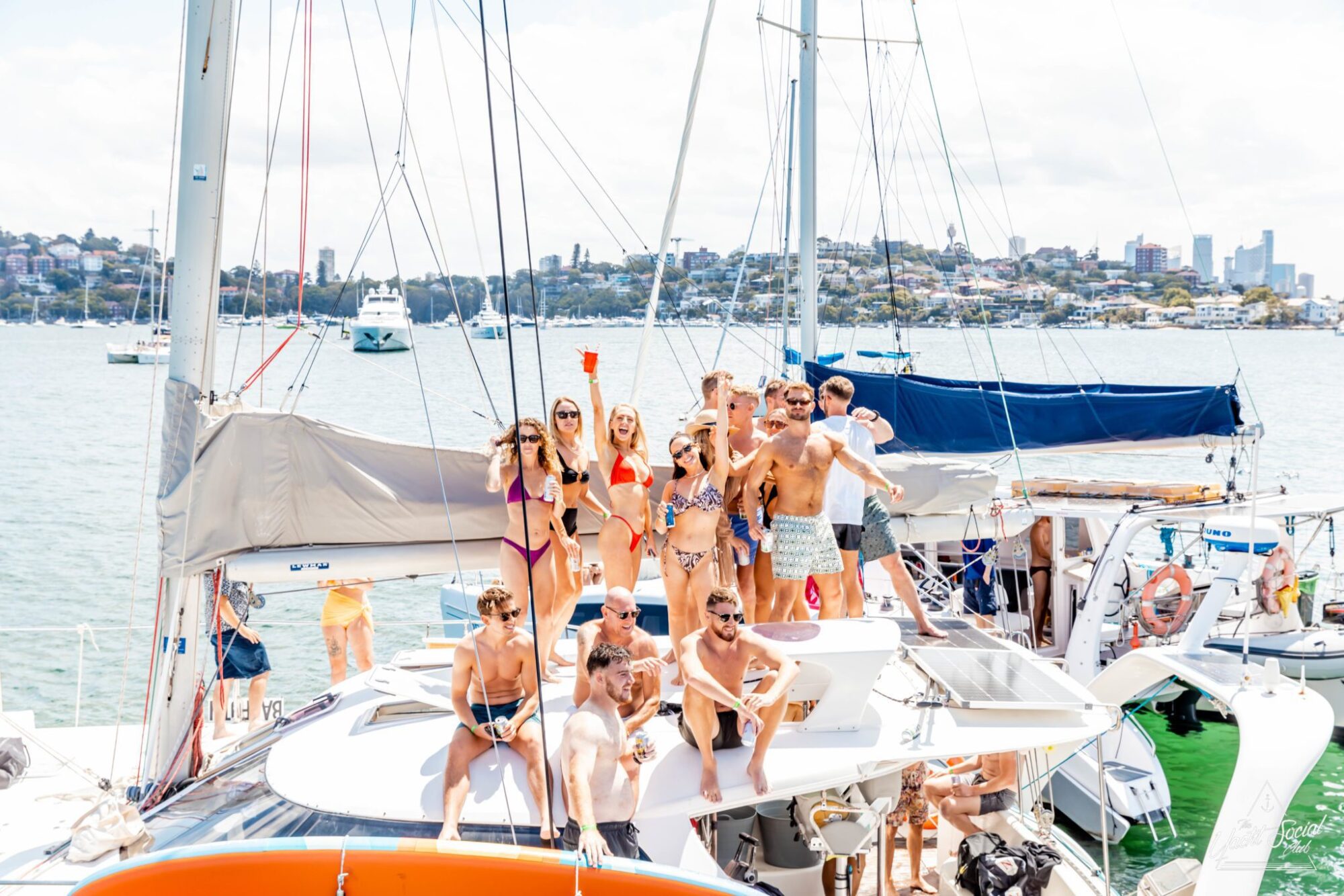 A group of people in swimwear stand and sit on a sailboat, some holding drinks, on a sunny day with other boats and a city skyline in the background. The scene epitomizes the lively atmosphere typical of catamaran parties in Sydney.