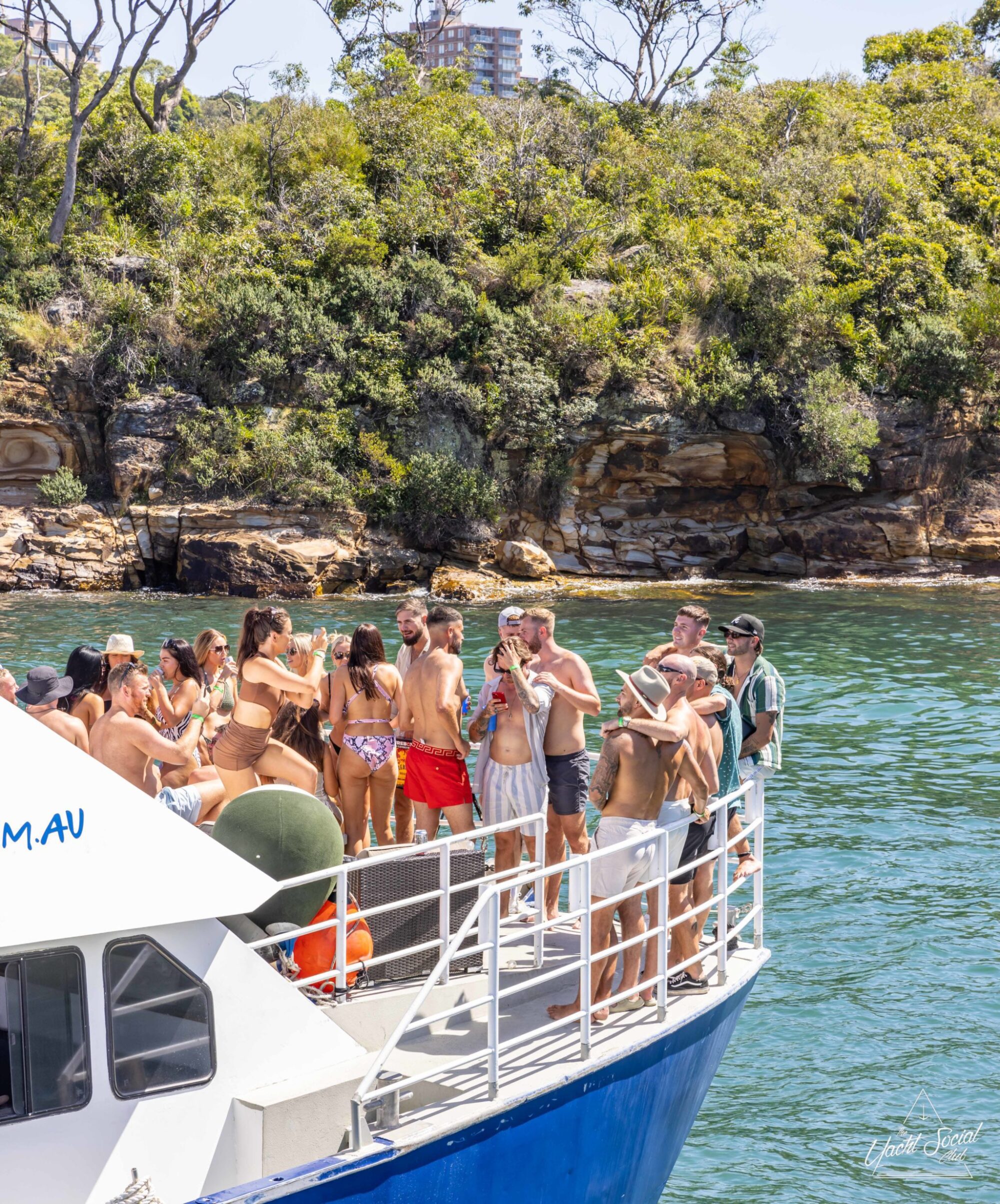 A group of people in swimwear is standing on a boat near a rocky shoreline with trees in the background, enjoying their time with a DJ boat hire Sydney.