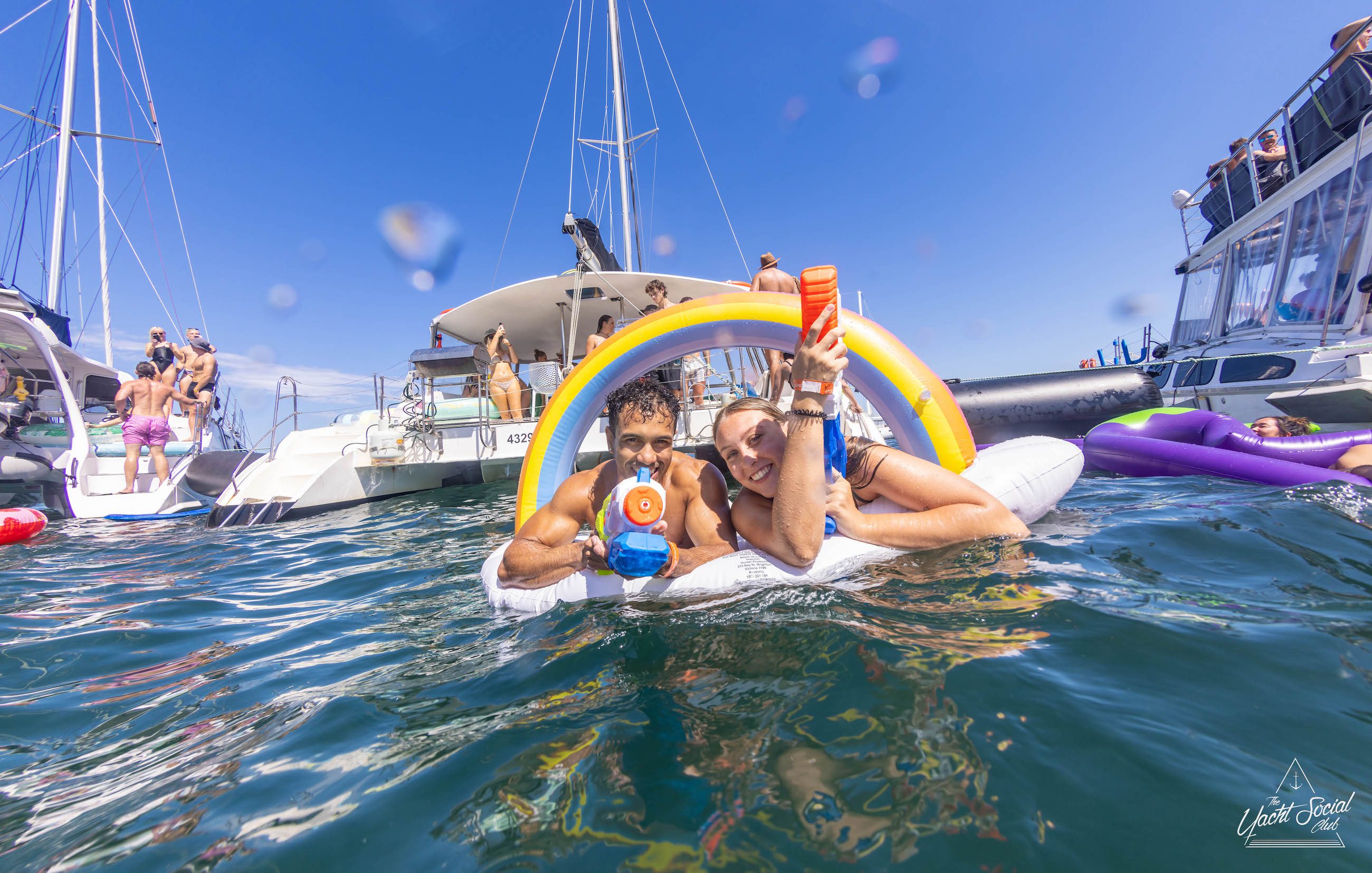 Two people float on an inflatable unicorn in the ocean, surrounded by boats and other people. They hold water guns and smile under a clear blue sky, enjoying what feels like a spontaneous Sydney boat party hire experience.