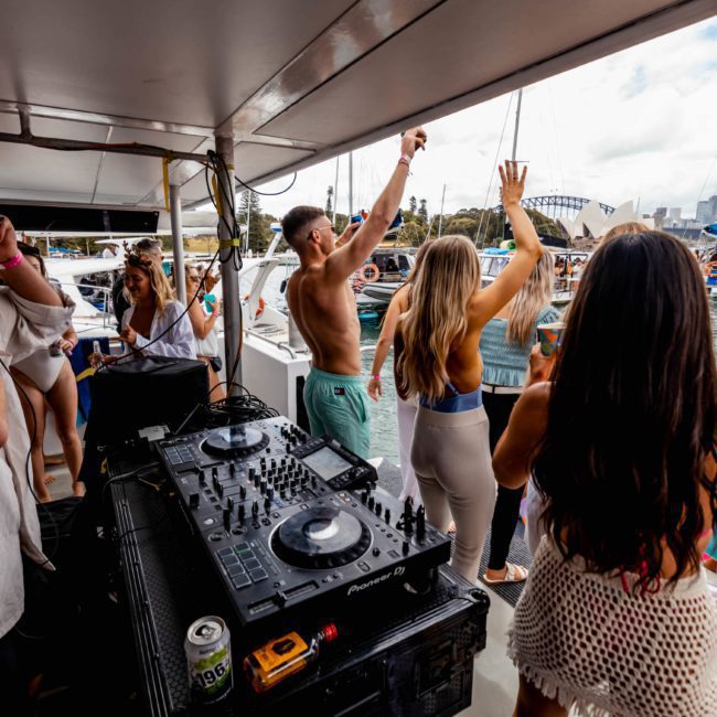 People on a private yacht charter in Sydney Harbour are dancing and enjoying a party near the water, with DJ equipment and inflatables in the background. The scene includes a clear day and views of nearby boats and structures.