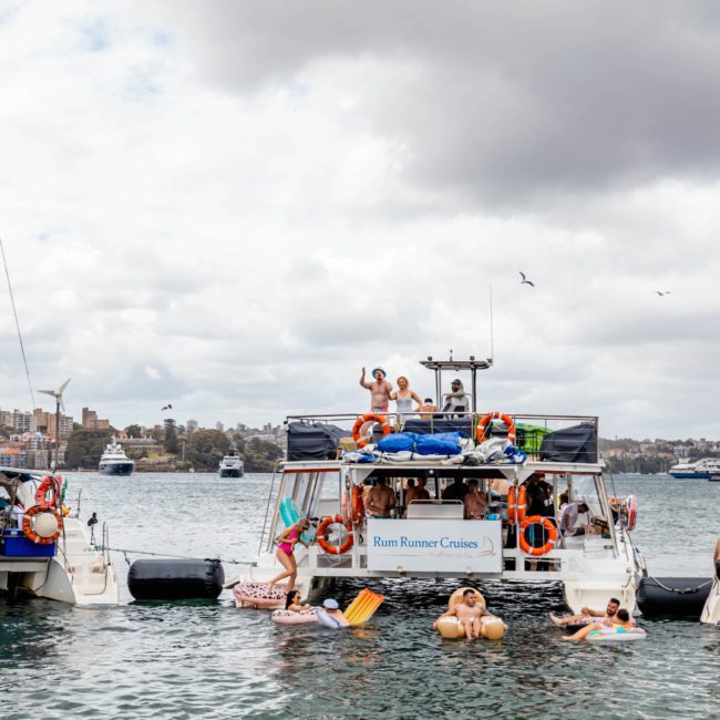 People are enjoying a lively gathering on boats and inflatables in a harbor, under a cloudy sky. Several boats, including luxury yachts, are anchored close together, with city buildings visible in the background.