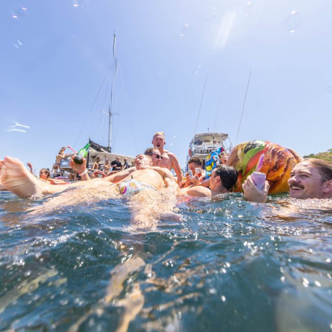 People are swimming and floating in a body of water near boats under a clear sky, enjoying a lively atmosphere thanks to the DJ boat hire Sydney. Bubbles float through the air, adding to the fun ambiance.