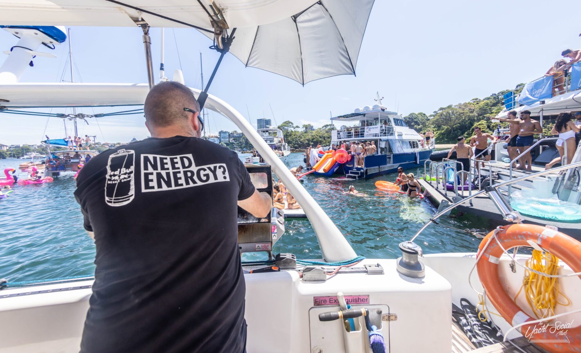 A man in a black "Need Energy?" shirt stands on a boat, facing a lively scene filled with people on inflatables, boats, and floating structures in a sunny, bustling waterway—perfect for those considering DJ boat hire Sydney.