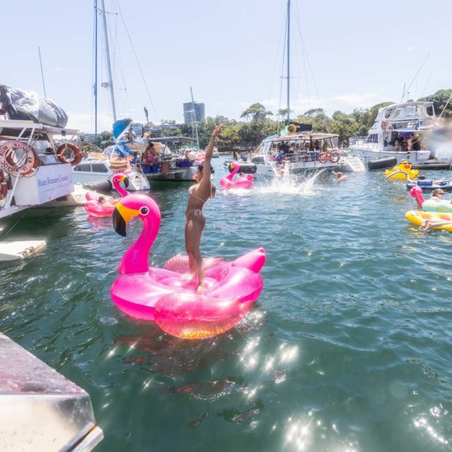 People enjoy a sunny day on a lake, with some floating on colorful inflatables, including a woman standing on a large pink flamingo float. Nearby, a few boats are docked, perfect for those considering private yacht charter Sydney Harbour for an added touch of relaxation and luxury.