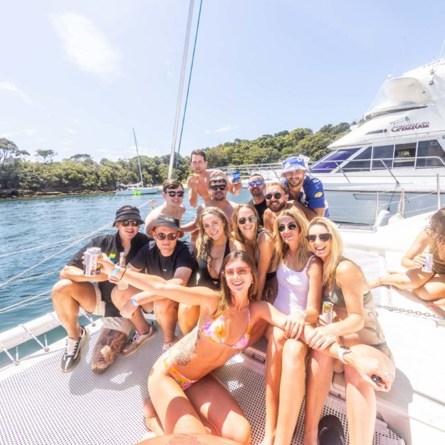 A group of people in swimwear are smiling and posing for a photo on the deck of a luxury yacht hire Sydney, with trees and a clear sky in the background.