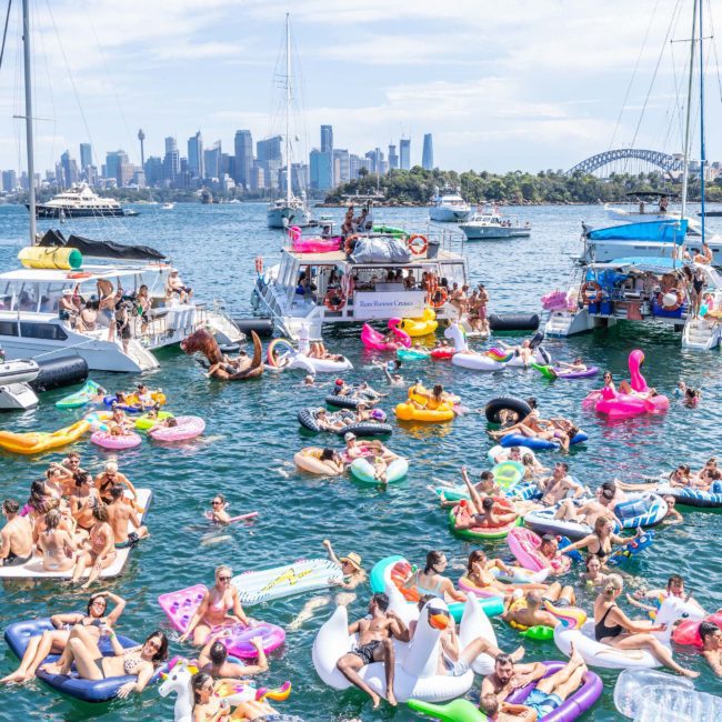 A large group of people on colorful inflatable floats in the water near boats, with a city skyline and the Sydney Harbour Bridge visible in the background. Luxury yacht hire Sydney enhances this vibrant scene, offering an elegant touch to your aquatic adventure.
