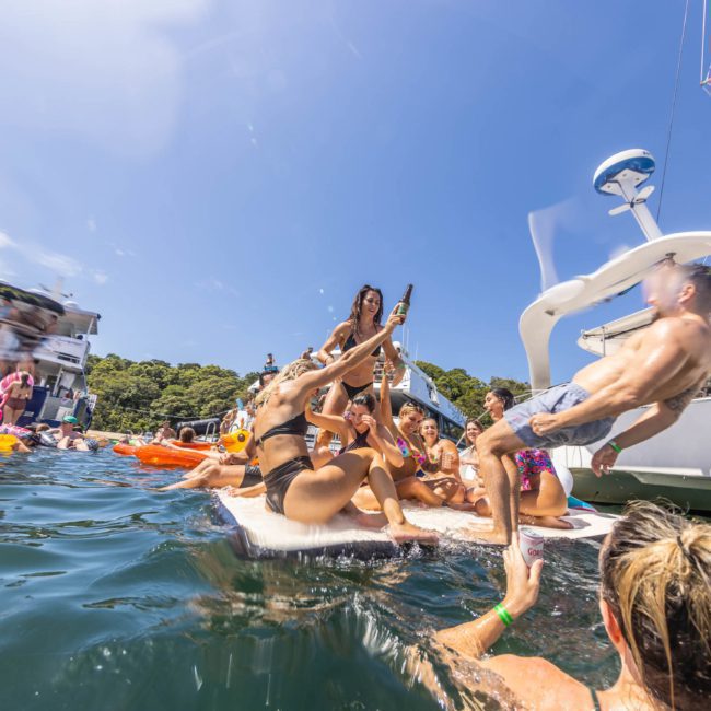 People enjoying a party on boats and inflatables in the water, with some individuals cheering and holding drinks under a clear sky. A luxury yacht hire in Sydney adds an elegant touch to the festivities.