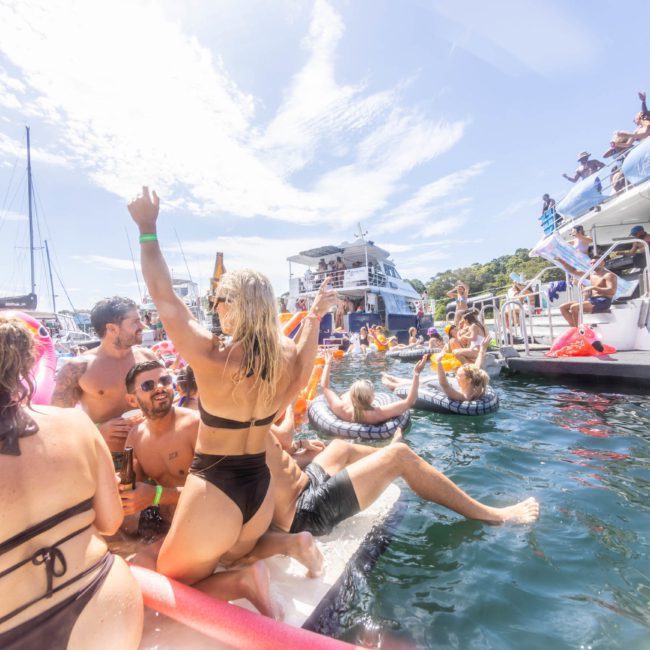 A large group of people in swimsuits enjoying a Catamaran party Sydney on the water with inflatables and music, surrounded by boats and a clear sky.