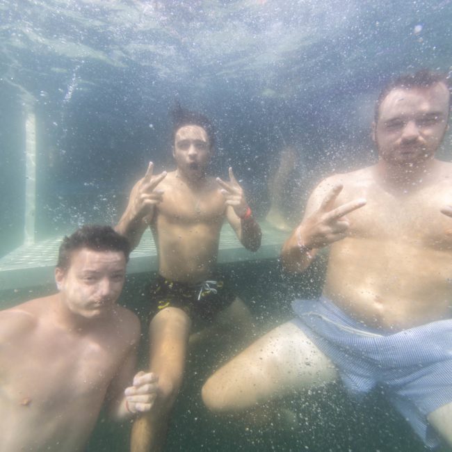 Three men underwater, each holding up peace signs with both hands. They are wearing swim trunks and appear relaxed, enjoying the unique vibe of a Sydney boat party hire.