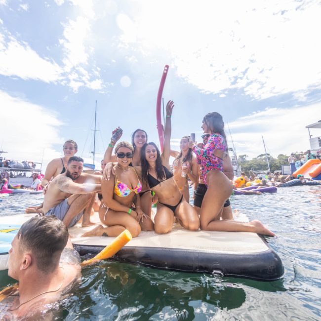 A group of people in swimsuits gather and smile on a floating platform in a body of water, with boats and more people in the background under a partly cloudy sky. It's the perfect scene for a catamaran party Sydney has to offer.
