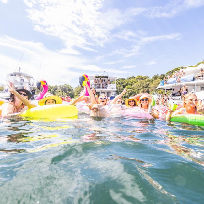 A group of people in colorful inflatable rings enjoying the water near several anchored boats on a sunny day, perfect for a Sydney boat party hire.