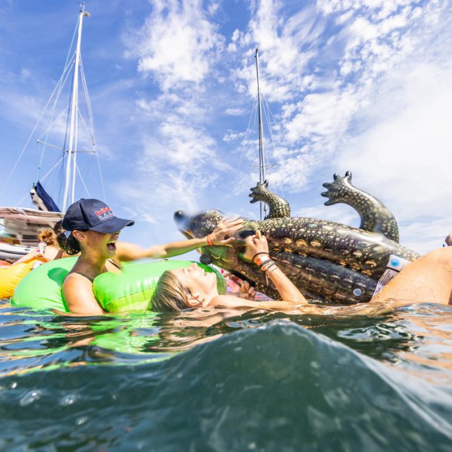 People in water with inflatable floats, including a green alligator and a black dragon. Boats and blue sky in the background, perfect for a Catamaran party Sydney.