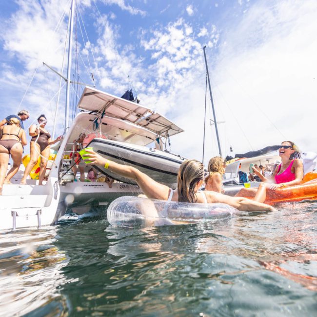 People in swimsuits enjoying the sea, some on floats, and others boarding or disembarking a catamaran party Sydney anchored under a sunny sky.