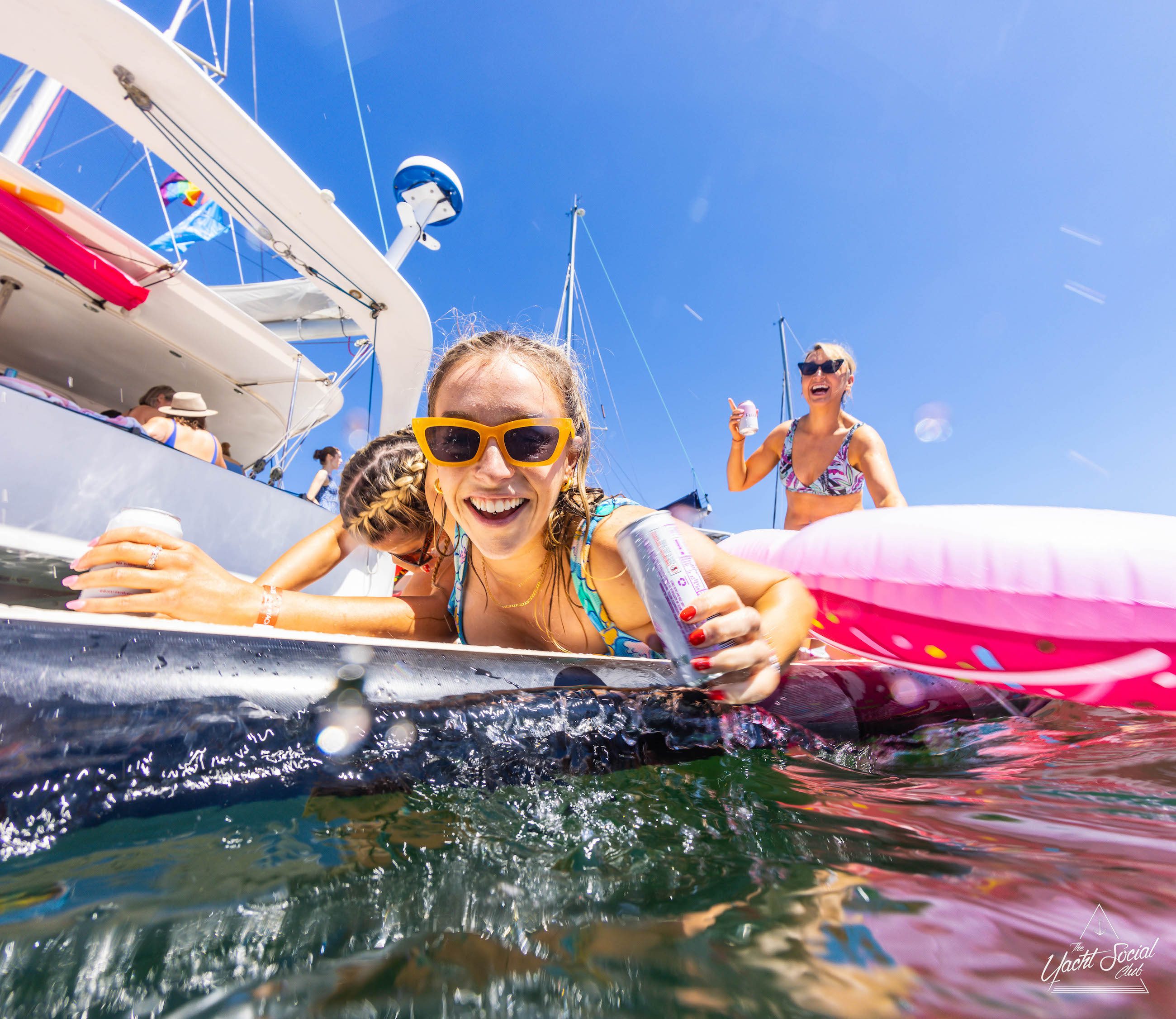 People enjoy a sunny day on a private yacht charter in Sydney Harbour, with one person in yellow sunglasses smiling at the camera and holding a can, while others in swimsuits relax on an inflatable float.