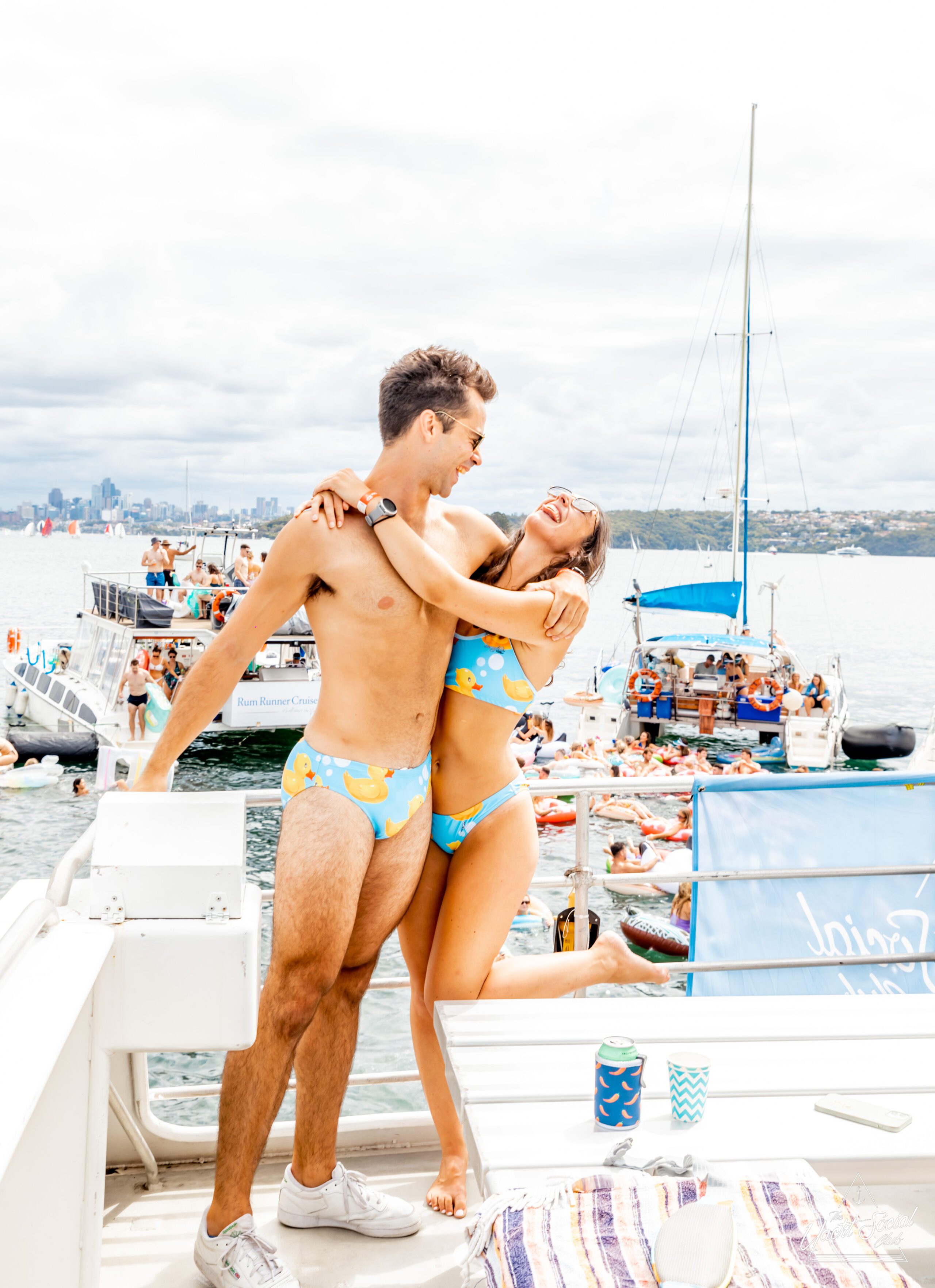 A man and woman, both in matching floral swimwear, smile and pose together on a boat deck during a lively Sydney boat party hire. The background shows other boats and people in the water under a cloudy sky.