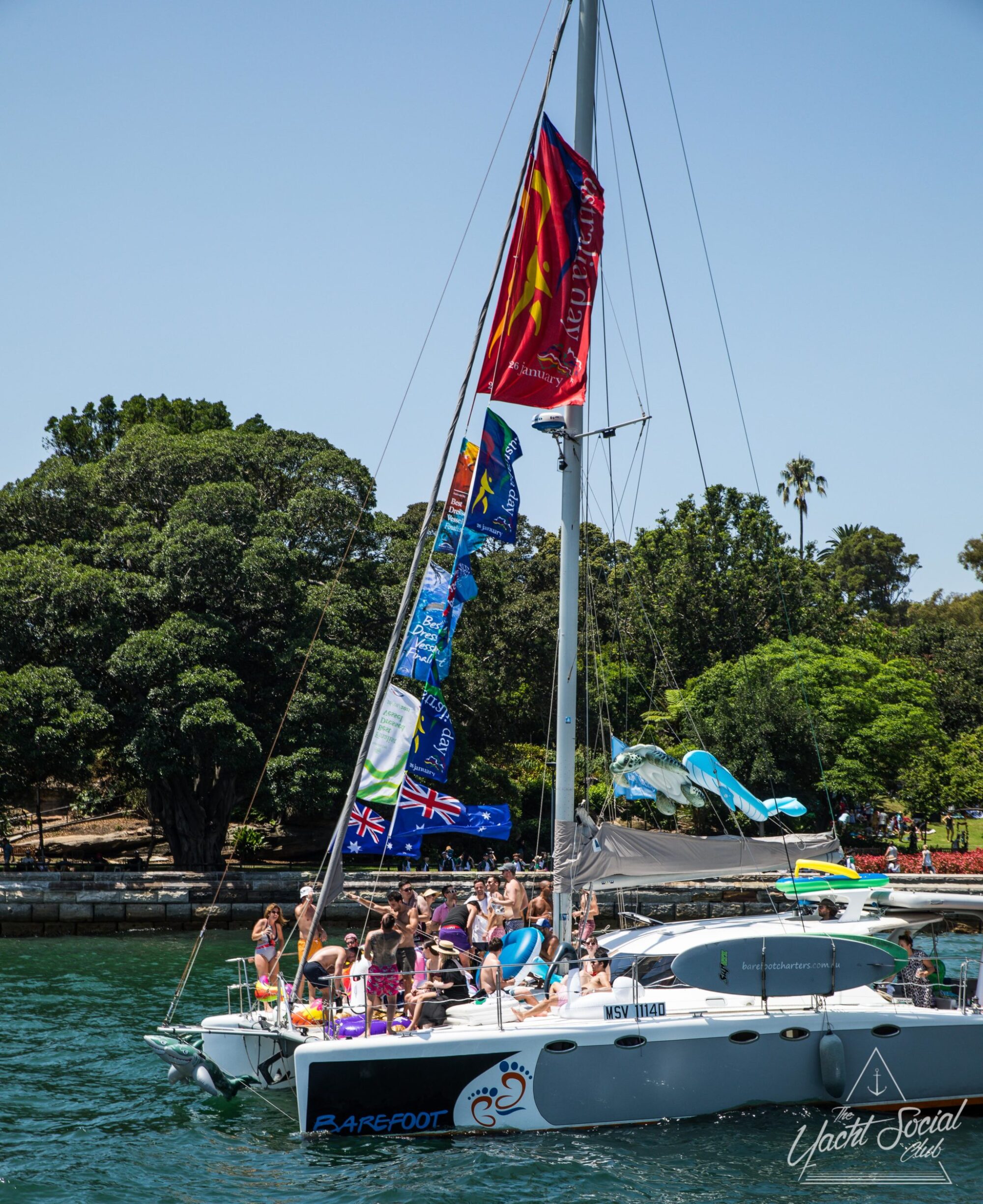 Sailboat with colorful flags, with a group of people onboard in swimwear, docked near a green park shoreline on a sunny day. Text on sail includes "Barefoot" and "Yacht Social." Ideal for those seeking luxury yacht hire Sydney or looking to experience a memorable Catamaran party Sydney event.