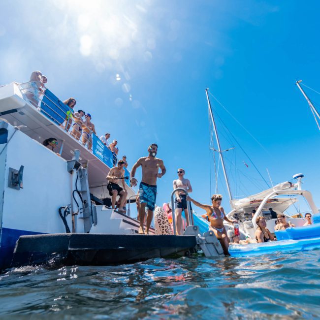 People on and around a luxury yacht anchored in the water under a clear blue sky. Some are on the boat's deck while others are in the water near an inflatable float. Several other boats are visible nearby, adding to the vibrant atmosphere of Sydney Harbour.
