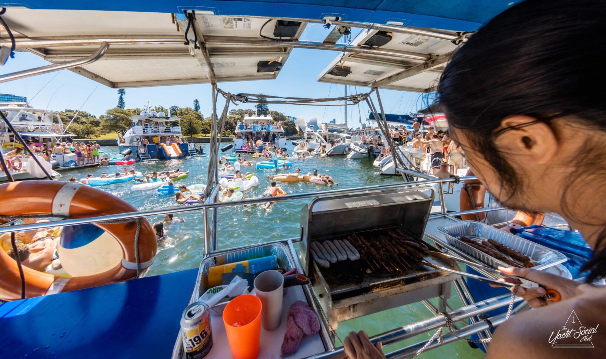 A person grills food on a boat's barbecue while a large group of people in swimwear enjoys the water and other boats in the background, typical of an exciting Sydney boat party hire.