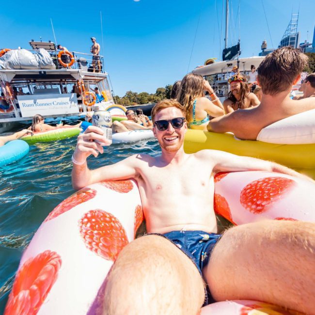 A man in sunglasses lounges on an inflatable in the water, holding a beverage. Other people are on inflatables and boats in the background, enjoying what seems like a private yacht charter in Sydney Harbour.