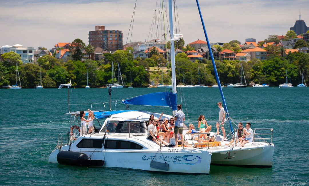 A group of people relax and socialize on a catamaran named Catalyst, anchored near a residential shoreline with greenery and buildings in the background. Enjoy the ultimate experience with Sydney boat party hire as they celebrate on this beautiful day.