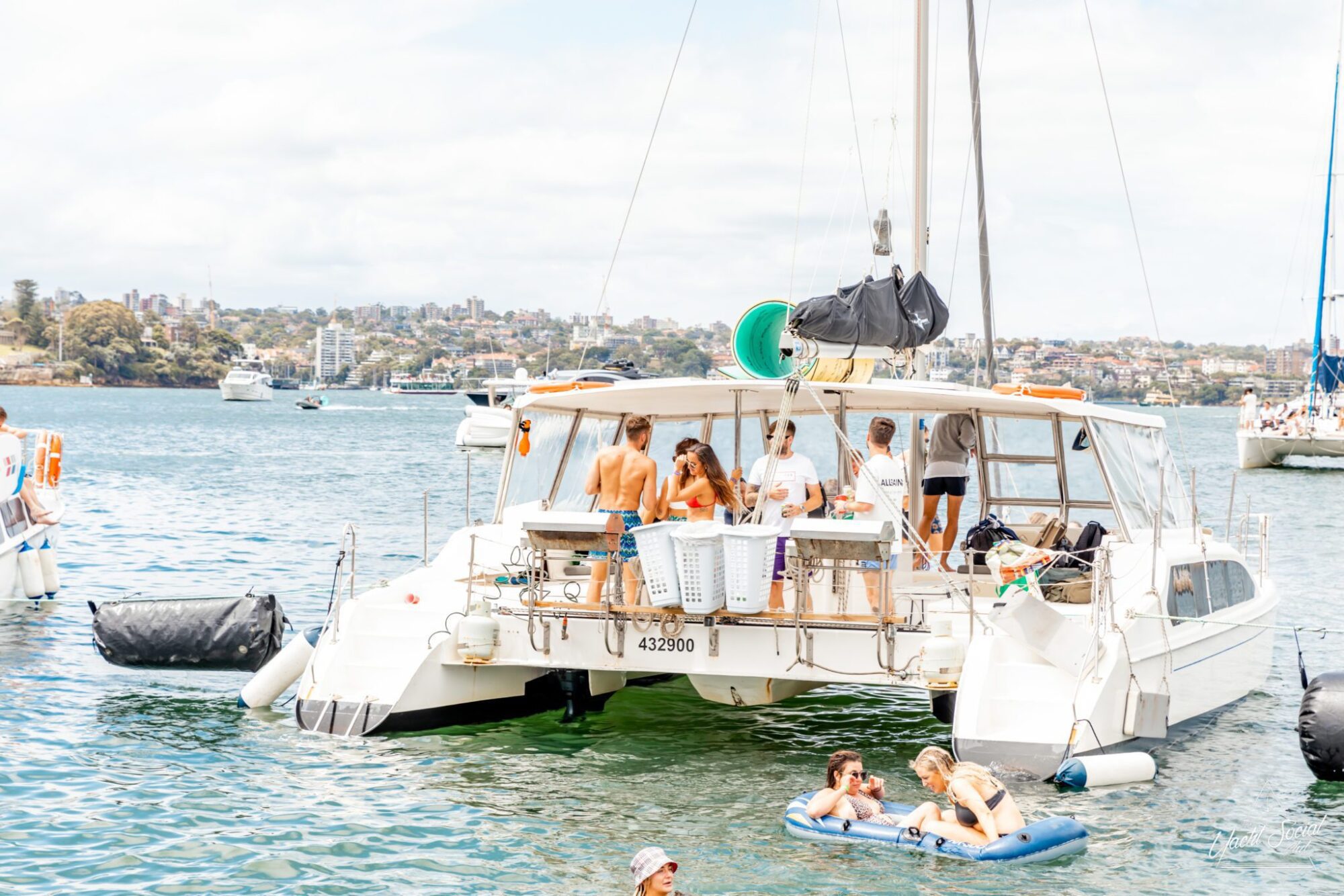 A group of people stands and interacts on a stationary catamaran in Sydney Harbour, enjoying a lively Catamaran party. Meanwhile, two individuals rest on an inflatable raft nearby.